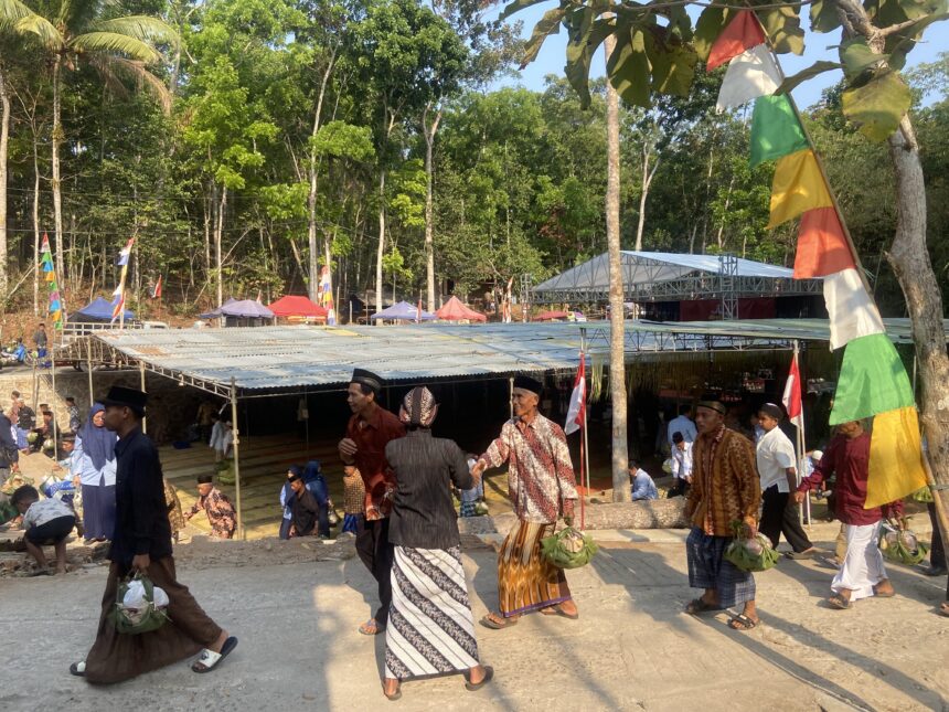 People in traditional clothing carrying woven baskets at an outdoor festival with colorful flags and stalls under trees.