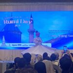 Large blue stage screen displaying 'Rumi Day 2025' with a crowd in front, many people recording with phones.