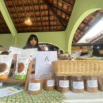 Market stall with jars of abon spices and printed menus on the counter; a woman stands behind the display under a green arched roof.