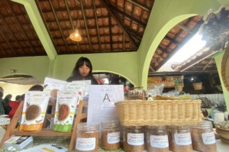 Market stall with jars of abon spices and printed menus on the counter; a woman stands behind the display under a green arched roof.