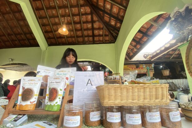 Market stall with jars of abon spices and printed menus on the counter; a woman stands behind the display under a green arched roof.