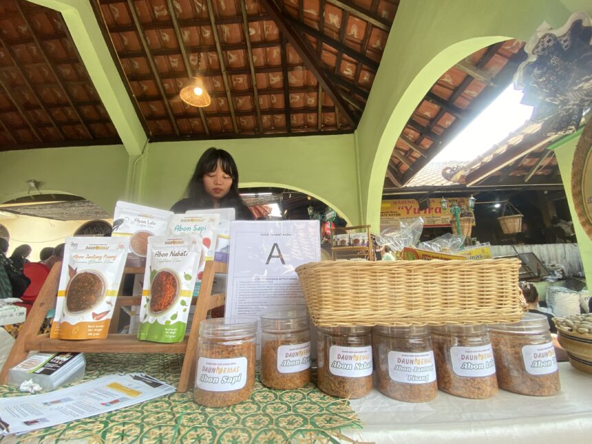 Market stall with jars of abon spices and printed menus on the counter; a woman stands behind the display under a green arched roof.