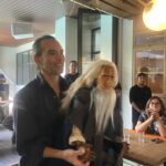 Man in a black shirt smiling at a casual indoor gathering, standing next to a person wearing a long white wig and mask.
