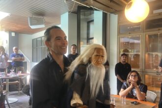 Man in a black shirt smiling at a casual indoor gathering, standing next to a person wearing a long white wig and mask.