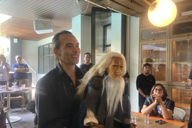 Man in a black shirt smiling at a casual indoor gathering, standing next to a person wearing a long white wig and mask.