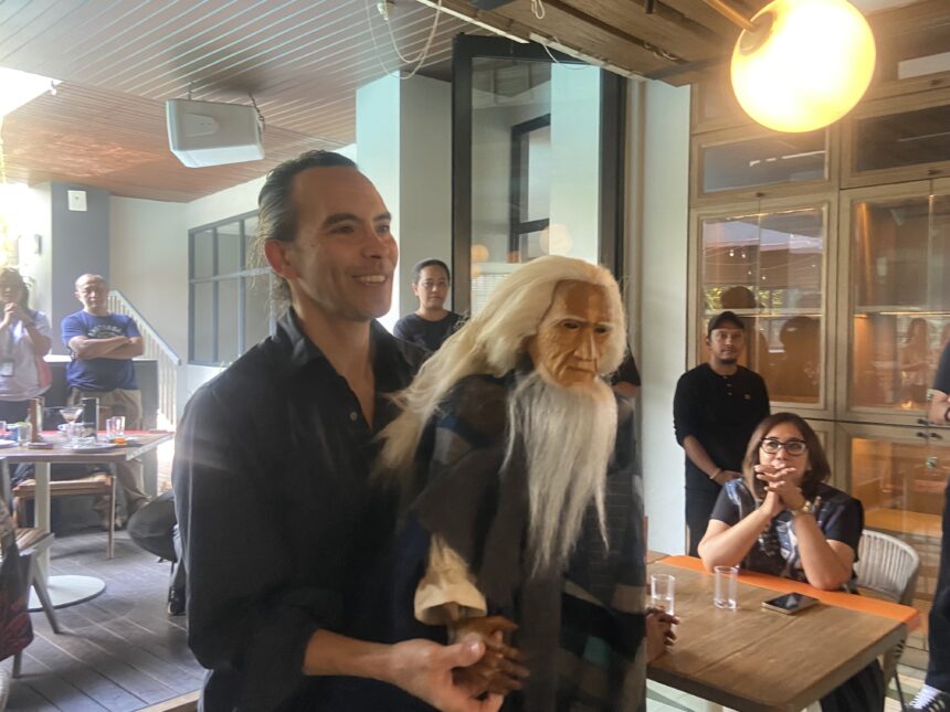 Man in a black shirt smiling at a casual indoor gathering, standing next to a person wearing a long white wig and mask.
