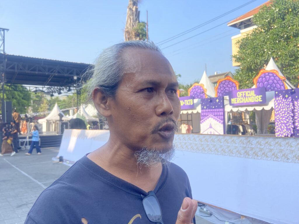 IMG_4892 - Mabur.co Close-up of a man with gray hair and stubble at an outdoor festival, with purple 'Official Merchandise' tents in the background.