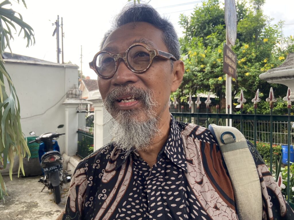 Smiling man with round glasses and gray beard, wearing a patterned shirt, standing outdoors with a backpack strap over his shoulder.