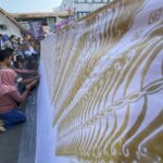 Crowd of people kneeling along a long decorative banner with gold patterns at an outdoor event.