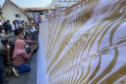 Crowd of people kneeling along a long decorative banner with gold patterns at an outdoor event.