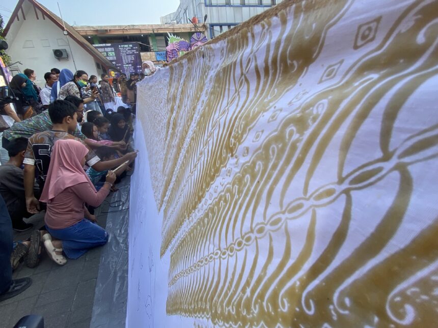 Crowd of people kneeling along a long decorative banner with gold patterns at an outdoor event.