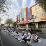 People praying on a long blue mat in the middle of a city street, with shoes lined up nearby and storefronts in the background (Dunkin’ sign visible).