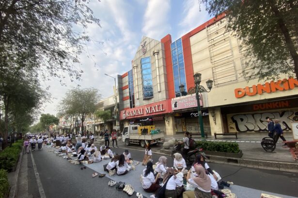 People praying on a long blue mat in the middle of a city street, with shoes lined up nearby and storefronts in the background (Dunkin’ sign visible).