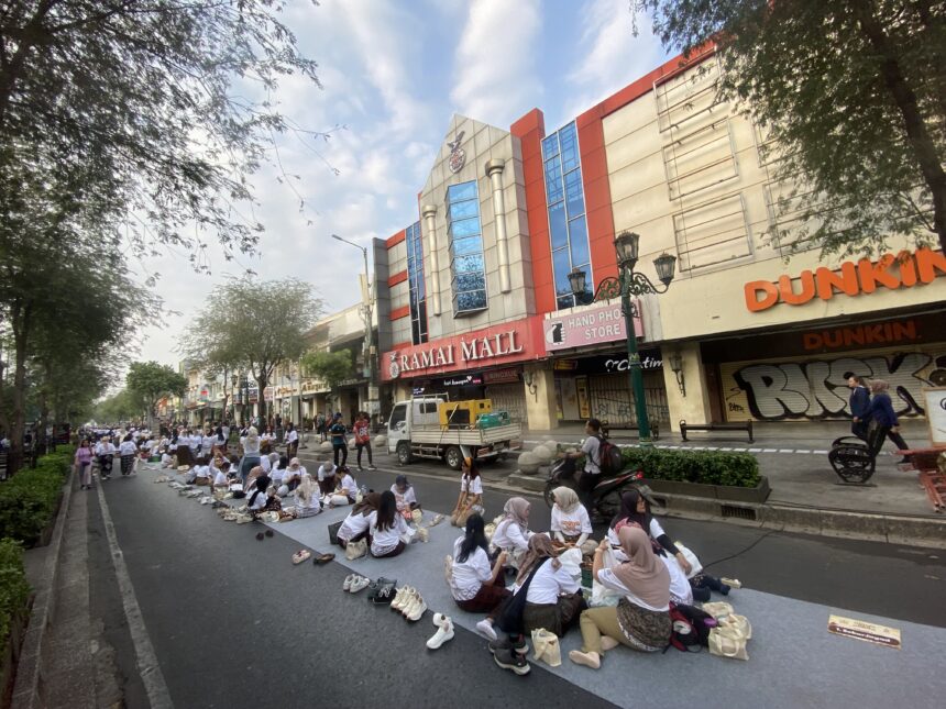 People praying on a long blue mat in the middle of a city street, with shoes lined up nearby and storefronts in the background (Dunkin’ sign visible).