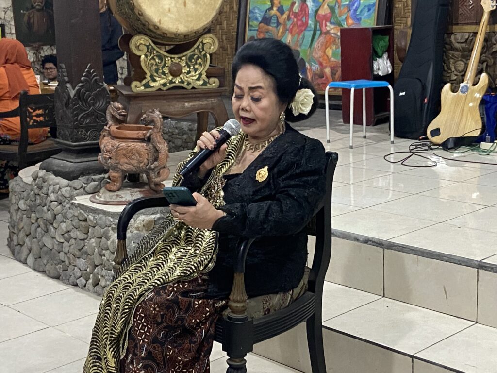 Older woman in a black and gold traditional outfit singing into a microphone while looking at a phone in a decorative indoor setting