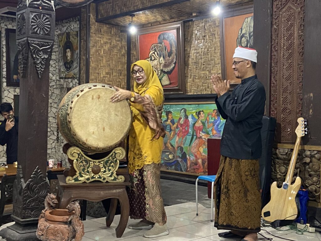 Woman in a yellow hijab plays a large traditional drum (kendang) while a man in a black outfit and white hat claps nearby in a culturally themed room.