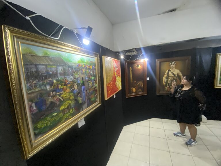 Woman in a black dress and sneakers taking a photo inside a small art gallery with black walls and white tiled floor, several framed paintings on display.