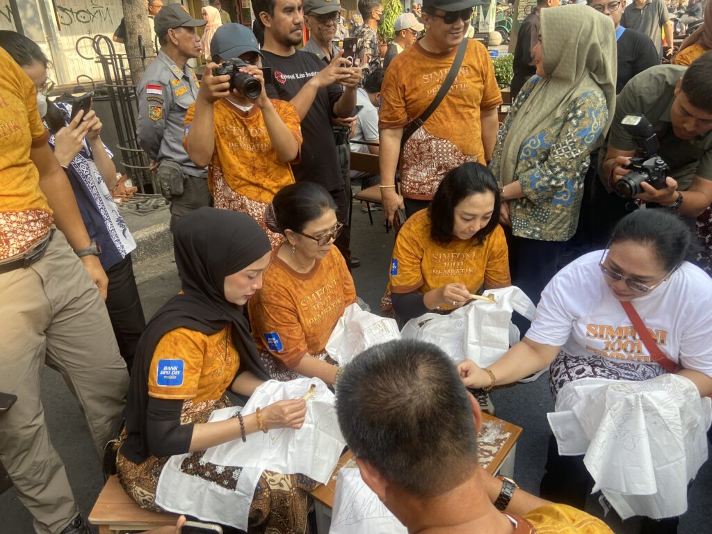 Crowded street scene with people in orange shirts signing papers at a table while photographers and bystanders take photos, some wearing hijabs.