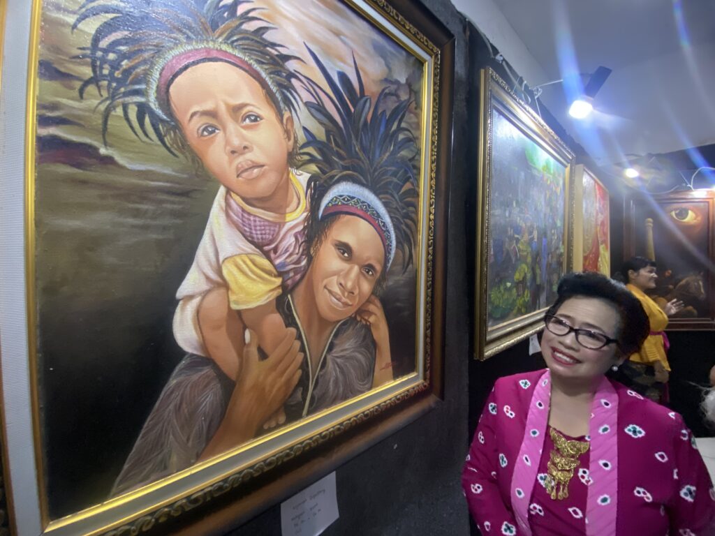 Smiling woman in a bright pink jacket stands beside a large framed painting of two Indigenous children with feathered headdresses.