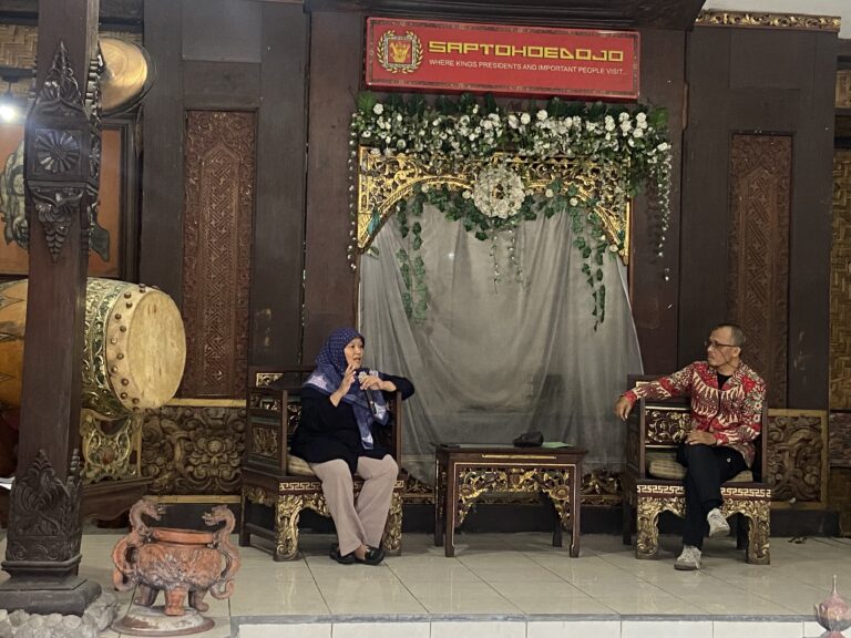 Two people sit on ornate carved chairs on a decorated stage; a large drum stands to the left and a flower garland arches over a draped backdrop with a red sign above.