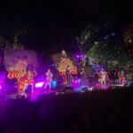 Group of performers in bright, patterned costumes on a lit outdoor stage at night, with purple stage lights and trees in the background.