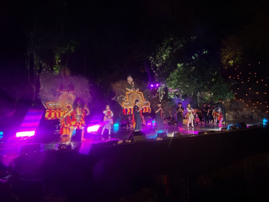 Group of performers in bright, patterned costumes on a lit outdoor stage at night, with purple stage lights and trees in the background.