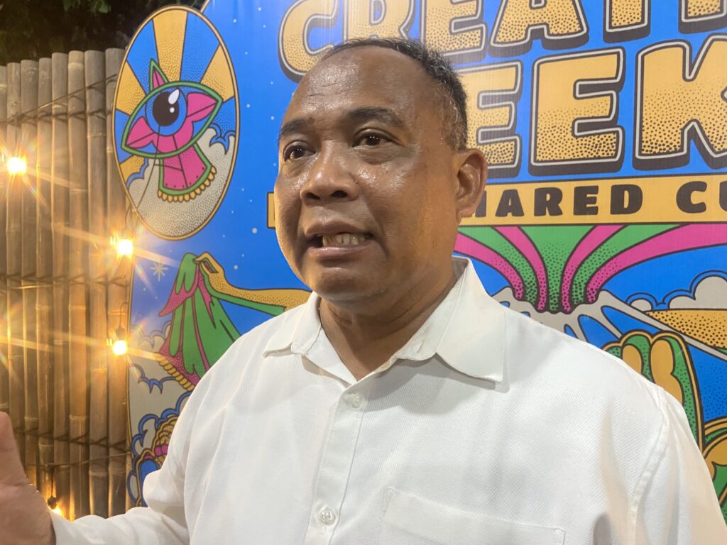 IMG_5053 - Mabur.co Close-up of a man in a white shirt taking a selfie in front of a colorful mural with bold graphics and text in yellow and blue.