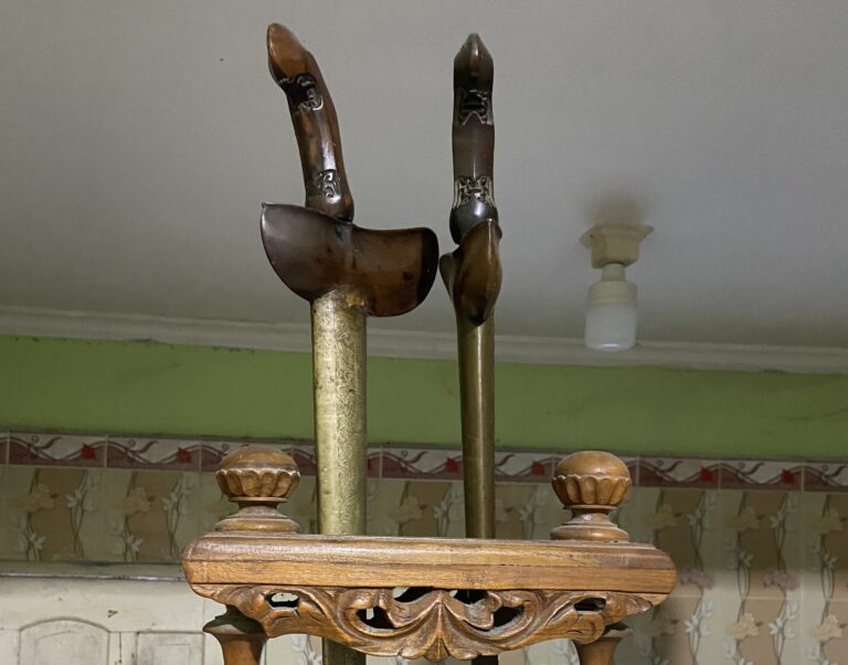 Antique wooden fireplace tool set on an ornate stand with tall tongs and brass columns in a vintage room.