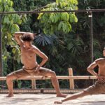 Two shirtless men practicing martial arts on a wooden platform outdoors, surrounded by lush green foliage and vines.