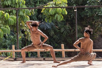 Two shirtless men practicing martial arts on a wooden platform outdoors, surrounded by lush green foliage and vines.