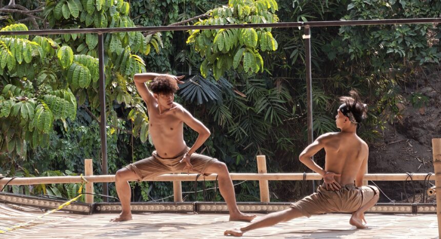 Two shirtless men practicing martial arts on a wooden platform outdoors, surrounded by lush green foliage and vines.