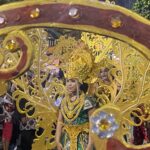 Parade participant in a gold ornate costume and headdress, framed by a decorative gold float, with spectators in back.