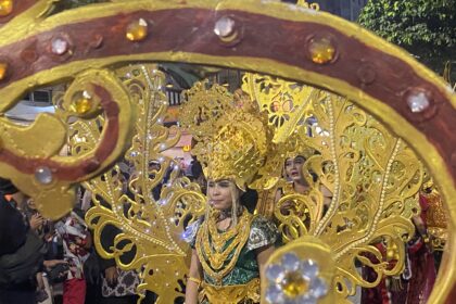 Parade participant in a gold ornate costume and headdress, framed by a decorative gold float, with spectators in back.