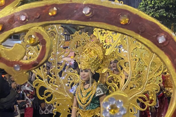Parade participant in a gold ornate costume and headdress, framed by a decorative gold float, with spectators in back.