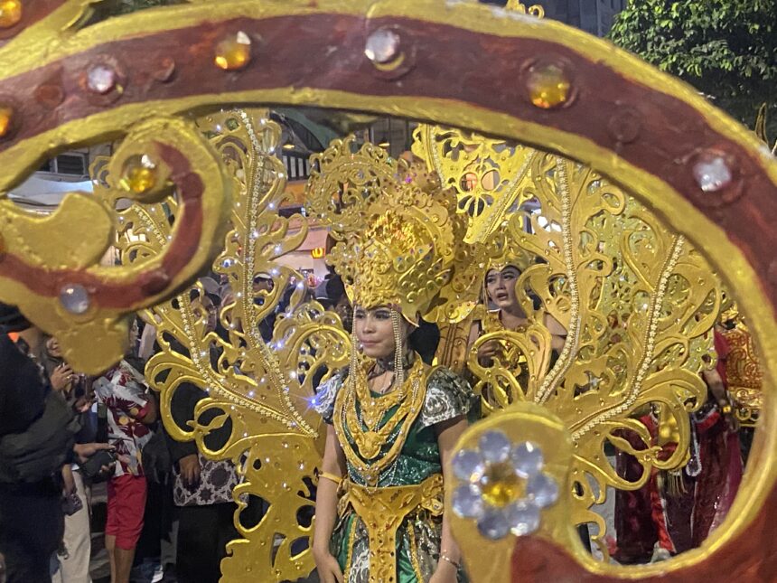 Parade participant in a gold ornate costume and headdress, framed by a decorative gold float, with spectators in back.