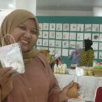 Smiling woman wearing a tan hijab and peach blouse holds up a white craft in a clear plastic bag at a craft fair, with artwork displayed on a blue-green wall behind her.