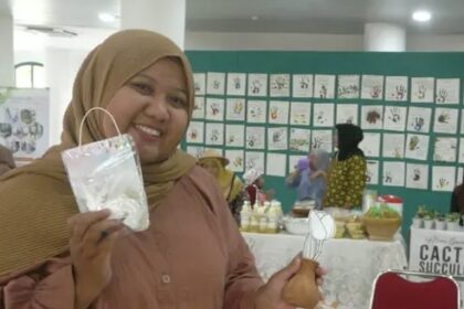 Smiling woman wearing a tan hijab and peach blouse holds up a white craft in a clear plastic bag at a craft fair, with artwork displayed on a blue-green wall behind her.