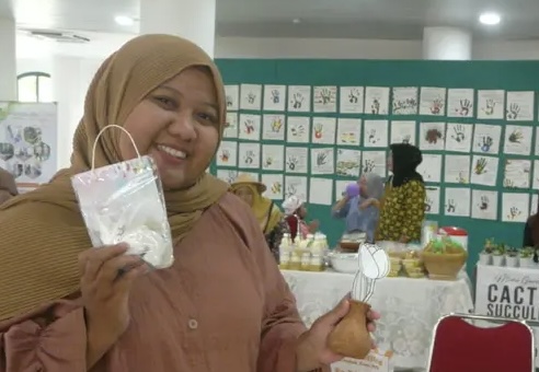 Smiling woman wearing a tan hijab and peach blouse holds up a white craft in a clear plastic bag at a craft fair, with artwork displayed on a blue-green wall behind her.