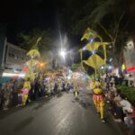 Nighttime street parade with dancers in bright yellow costumes marching down a crowded avenue, spectators lining both sides under festive lights.