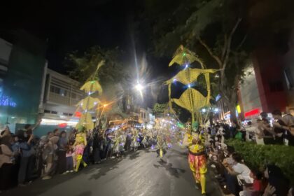 Nighttime street parade with dancers in bright yellow costumes marching down a crowded avenue, spectators lining both sides under festive lights.