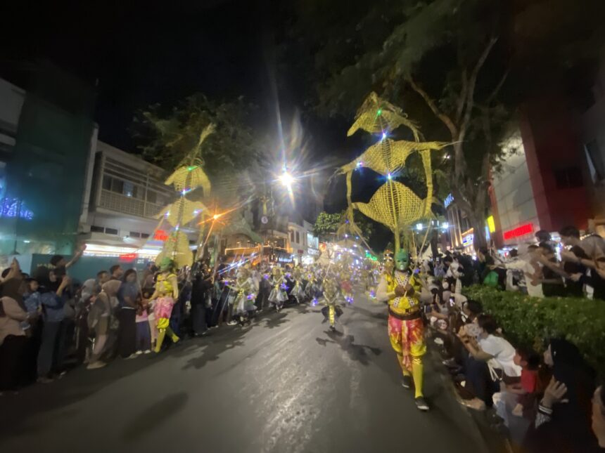 Nighttime street parade with dancers in bright yellow costumes marching down a crowded avenue, spectators lining both sides under festive lights.