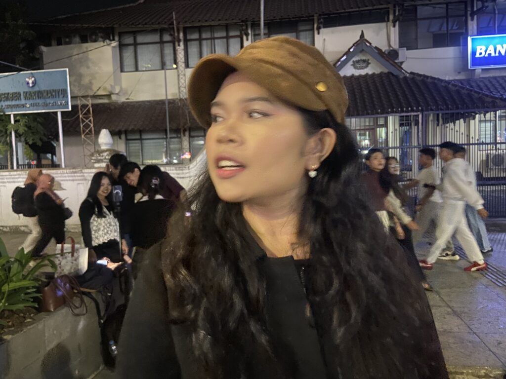 IMG_5293 - Mabur.co Close-up of a woman with long dark hair wearing a brown beret and black outfit, standing on a busy street at night with people and buildings in the background.