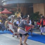 Group of Indonesian dancers in batik skirts and headdresses performing on a blue mat at an outdoor festival.