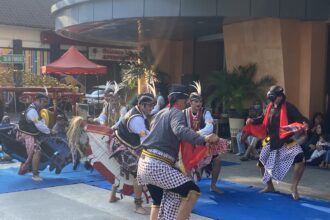 Group of Indonesian dancers in batik skirts and headdresses performing on a blue mat at an outdoor festival.