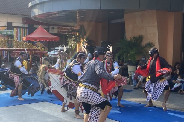 Group of Indonesian dancers in batik skirts and headdresses performing on a blue mat at an outdoor festival.