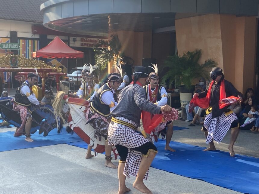 Group of Indonesian dancers in batik skirts and headdresses performing on a blue mat at an outdoor festival.