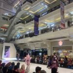 Fashion show on a glossy runway inside a decorated mall, models in pink traditional outfits walk as a seated audience watches from both sides.