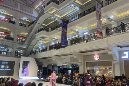 Fashion show on a glossy runway inside a decorated mall, models in pink traditional outfits walk as a seated audience watches from both sides.