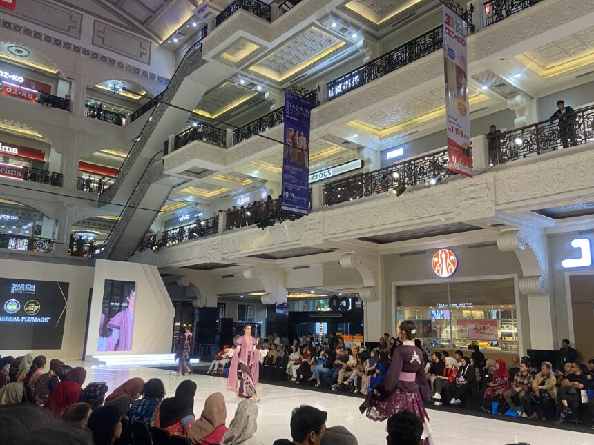 Fashion show on a glossy runway inside a decorated mall, models in pink traditional outfits walk as a seated audience watches from both sides.