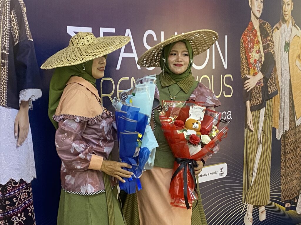 Two women in wide-brim straw hats and traditional outfits pose with flower bouquets at a fashion event backdrop in a purple-toned setting.